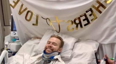 弗朗茨·沃尔躺在医院的病床上，脖子上戴着支架，上面写着“为爱干杯”的牌子